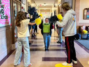 A student smiles as they pass through two lines of students cheering them on with pompoms. 