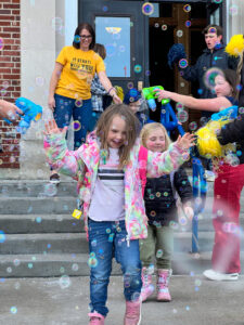 Elementary students show joy while older students blow bubbles at them. 